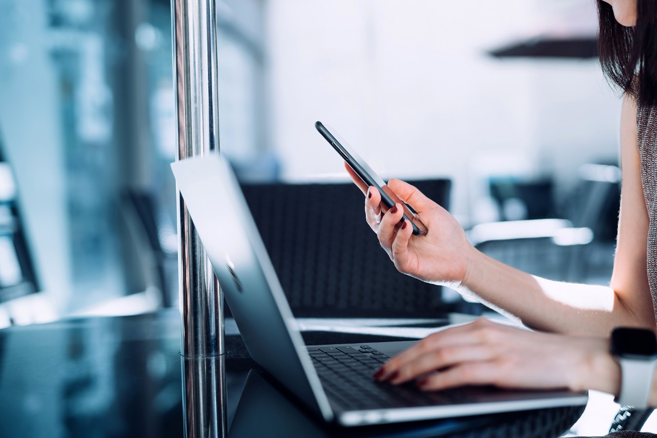 Cropped shot of young Asian businesswoman using smartphone and typing on keyboard on laptop on the go in sidewalk cafe. Remote working concept with flexible lifestyle. Business on the go